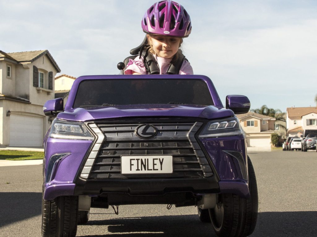 A little girl wearing a purple helmet and sitting in a purple toy car. Nameplate says "Finley" and there are houses in the background.