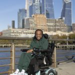 A man wearing a green hoodie and sweatpants sitting in a black wheelchair. There are skyscrapers in the background.