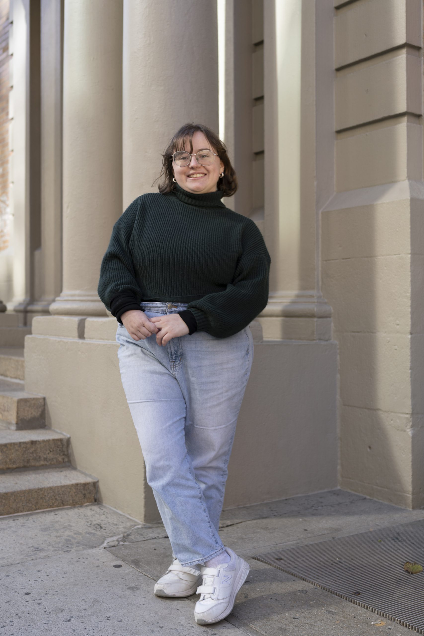 Full body photo of a smiling woman wearing a black turtleneck, blue jeans, and white shoes.