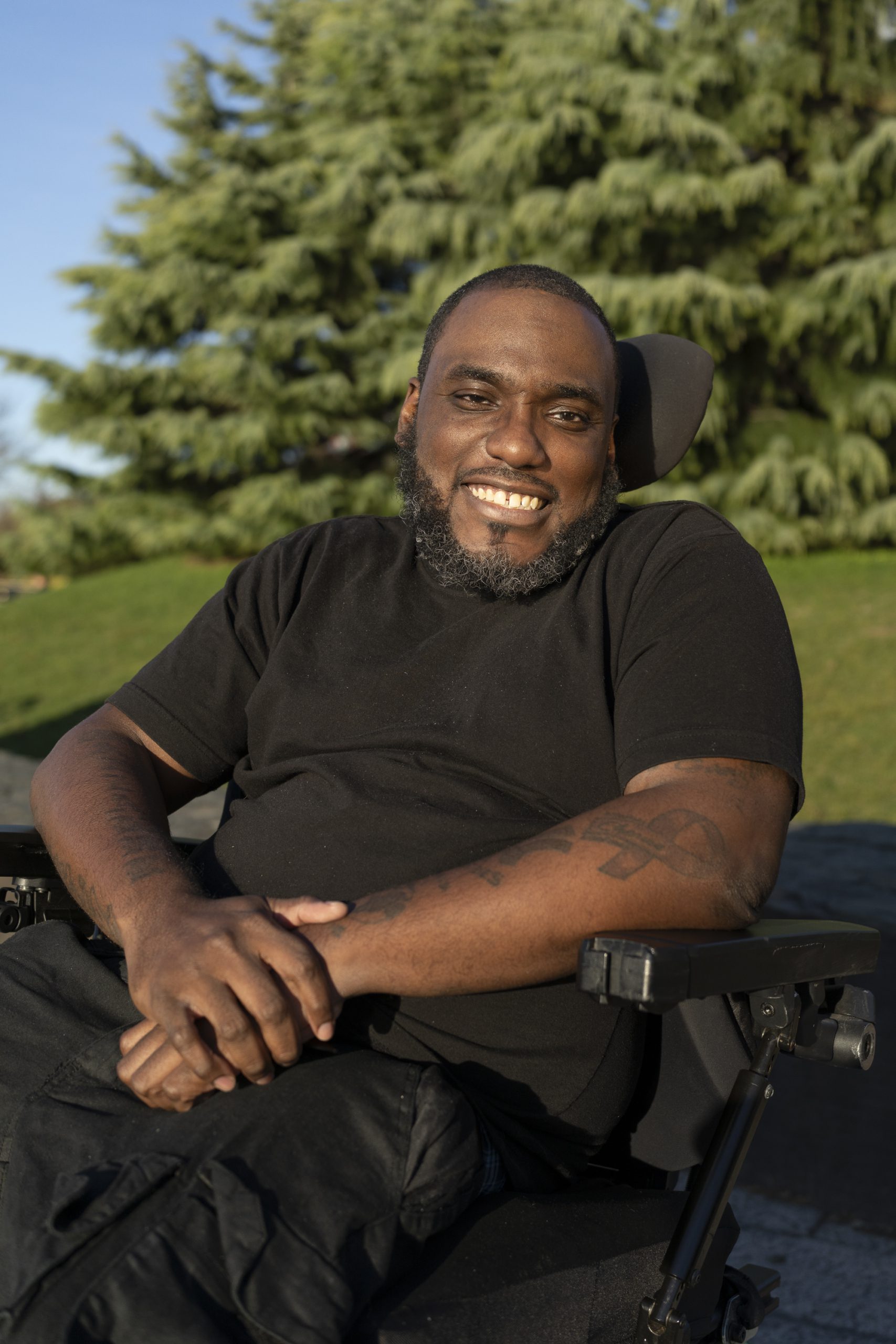 A man sitting in a wheelchair and smiling at the camera, with one hand over another. There are green trees in the background.