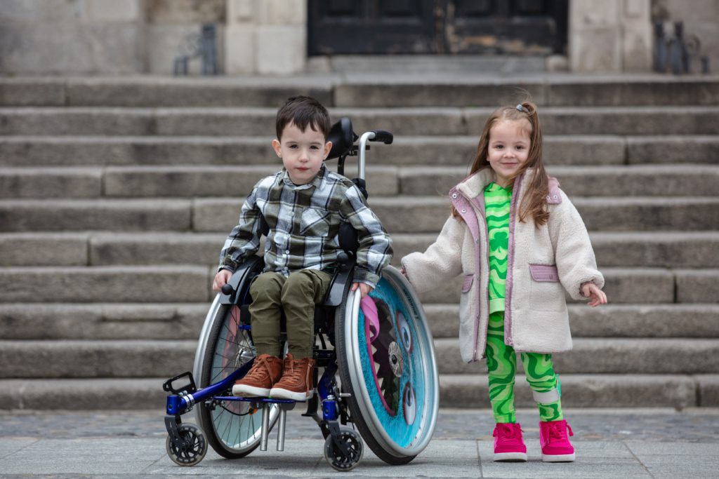 Smiling little girl standing next to little boy in wheelchair with steps in the background